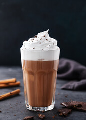 Hot chocolate with whipped cream and cinnamon in a tall glass on a dark background. Front view and copy space