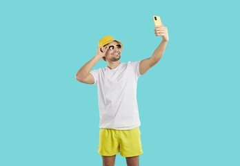 Wall Mural - Cheerful young man with mobile phone in his hand taking aselfie during summer sea vacation. Smiling man in summer shorts, T-shirt, panama hat and sunglasses is photographed on light blue background.