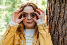 Smiling Woman Covering Eyes With Pine Cone In Forest