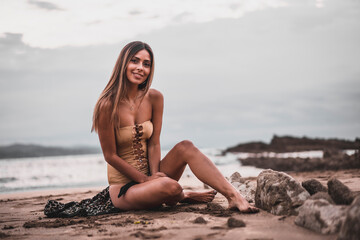 Wall Mural - Fashion posing of a brunette woman in a swimsuit and a pareo on the beach in summer, sitting on the sand