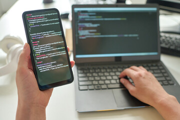 Close-up of programmer holding smartphone with codes in her hands and connecting it with laptop at table