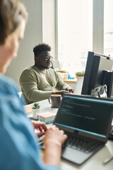 African young man sitting at table in front of computer monitor and typing with his colleague sitting opposite him and working on laptop