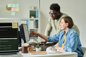 Young woman pointing at computer monitor and discussing new software together with her colleague during teamwork at office
