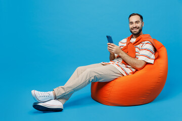 Wall Mural - Full body young smiling fun happy man 20s in orange striped t-shirt sit in bag chair hold in hand use mobile cell phone look camera isolated on plain blue background studio. People lifestyle concept