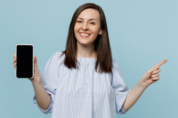 Wall Mural - Young fun woman she 20s in casual blouse hold in hand use mobile cell phone browsing internet with blank screen workspace area point index finger aside isolated on pastel plain light blue background