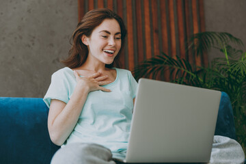 Young happy exultant fun woman she in mint t-shirt hold use work on laptop pc computer spread hands study online sit on blue sofa indoor rest at home in own room apartment. People lifestyle concept.