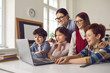 © Studio Romantic - Young smiling female teacher and pupils group watching funny educational video together on laptop at school classroom during lesson. Education in small group. Online meeting with classmates abroad