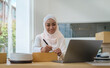 © wattana - Smiling young muslim woman  small business owner writing address on cardboard box, working at home office. Startup small business, Online selling, E-commerce.