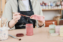 Young Creative Woman In Apron Holding Paintbrush And Clay Bowl Over Table While Coloring Handmade Earthenware In Marsala
