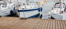 White Modern Sailboat (for Rent And Sale) Moored To A Pier In A Yacht Marina. Wooden Teak Deck. Nautical Vessel, Transportation, Amateur Sailing, Vacations, Cruising, Recreation Concepts