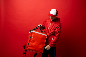 Sticker - Professional delivery employee man 20s in red cap T-shirt uniform workwear work as dealer courier with red thermal food bag backpack isolated on red background. Service concept.