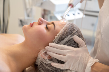 Wall Mural - Beautician applying electric Darsonval facial massage treatment with special tool over female patients forehead in beauty salon