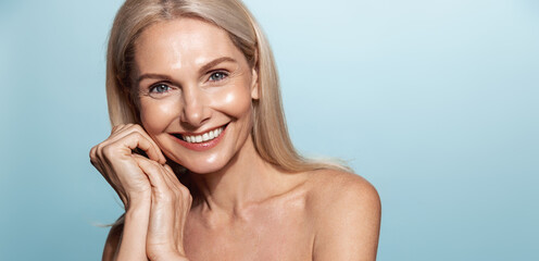 Elegant middle aged woman, head and shoulders with glowing skin, nourished, moisturized face and body, smiling at camera, blue background