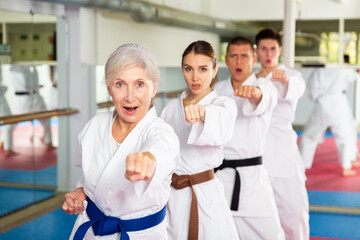 Wall Mural - Focused elderly woman wearing kimono standing in attacking stance, practicing movements of close combat punches in training room during group martial arts workout..