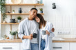 © Kateryna - Joyful positive hispanic man and caucasian woman, happy couple in love, stand at home in the kitchen, hug, drink tea or coffee together, enjoy spending time with each other, smile