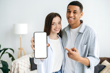 Positive modern caucasian girl and hispanic guy, a couple in love, in stylish clothes, stand in a cozy living room, show a smartphone with a blank white mockup screen, empty space for advertisement