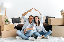 New Home. Happy Multiracial Young Couple In Love Sitting On The Floor In The Living Room Of Their New Home Between Cardboard Boxes, Making An Imaginary Roof With Their Hands, Looking At Camera, Smile