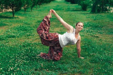 woman in a white tank top and multi-colored pants is doing yoga on the green grass in the city