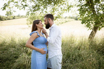 Wall Mural - young couple in love standing in high flower meadow in summer and cuddling and the woman is pregnant and has a blue dress on and the man has a full beard