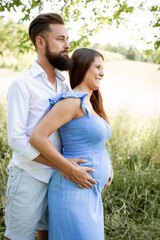 Wall Mural - young couple in love standing in high flower meadow in summer and cuddling and the woman is pregnant and has a blue dress on and the man has a full beard