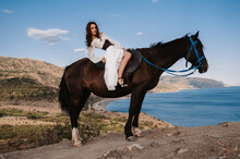 A Young Lady Rider In A White Dress Poses In The Saddle On A Horse Leaning Back Against A Beautiful Mountain Landscape. The Concept Of Romance, Fairy Tales. Example Of A Finished Book Cover