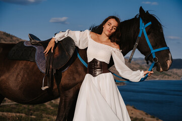 A young girl rider poses next to her horse against the backdrop of a mountain and sea landscape. The concept of riding. Artistic photography. Ready-made cover for books and magazines.