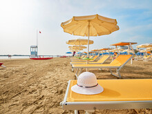 White Straw Hat On A Beach Bed With Colorful Umbrellas On A Sunny Summer Day. In The Background A Lifeboat Alongside A Lifeguard Tower On The Shore Of The Adriatic Sea, Italy.