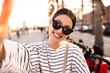 © Look! - Close-up of young caucasian woman in sunglasses smiling taking selfie in street. Brunette wears striped sweatshirt for walk. People sincere emotions lifestyle concept.