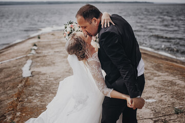 Groom in a black suit with a black bow tie with a beautiful bride in a white long dress embrace and kiss, holding hands near the river. Wedding portrait of newlyweds.