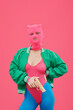 Portrait of fashion model in pink balaclava and body posing with gun against pink background expressing protest