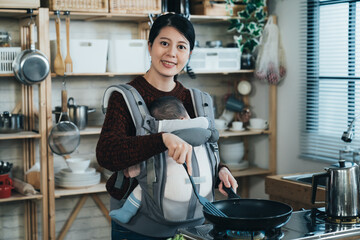 portrait cheerful asian housewife holding a spatula with infant child in the carrier is smiling at camera while preparing meal in the morning at home kitchen.