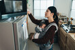 © PR Image Factory - portrait asian housewife with an infant baby in the carrier is putting plate into microwave oven to heat it up while preparing breakfast in the kitchen at home