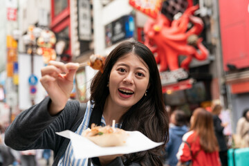 smiling Asian female sight-seer holding showing a takoyaki ball on pick and looking at camera while visiting shinsaibashi suji and doutonbori downtown Osaka japan