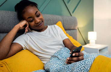 Wall Mural - Young African American woman laying in bed in the bedroom using smartphone.