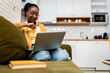 © Zoran Zeremski - Young African American woman sitting on sofa at home studying using laptop.
