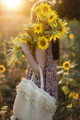 Wall Mural - Stylish young female in floral dress walking with sunflowers in warm sunset light in summer field. Tranquil atmospheric moment in countryside. Beautiful woman holding sunflowers in evening meadow