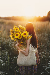 Wall Mural - Beautiful woman holding sunflowers in warm sunset light in meadow. Tranquil atmospheric moment in countryside. Stylish young female in floral dress walking with sunflowers in summer evening field