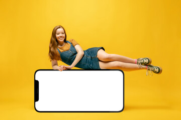 Young girl lying on huge 3d model of cellphone with blank white screen isolated on yellow background, Youth, new app or website, copy space for your ad, mockup