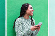 © VISTA by Westend61 - Happy young woman with smart phone standing in front of green wall