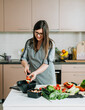 © Tijana - Smiling Woman in Apron Putting Sliced Cucumber and Tomato in a Salad Bowl on a Kitchen Desk