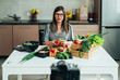 © Tijana - Serious Woman Holding Radish While Sitting At Kitchen Desk And Filming Video For Her Food Channel