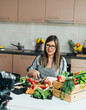 © Tijana - Smiling Woman Cutting Vegetables While Sitting At Kitchen Desk And Filming Video For Her Food Channel