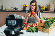 © Tijana - Happy Woman Holding A Tomato While Sitting At Kitchen Desk And Filming Video For Her Food Channel