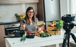 © Tijana - Happy Woman Holding Avocado And Zucchini While Sitting At Kitchen Desk And Filming Video For Her Food Channel