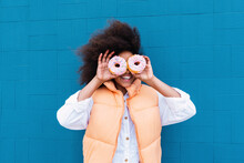 Happy Girl Covering Eyes With Doughnuts Standing In Front Of Blue Wall