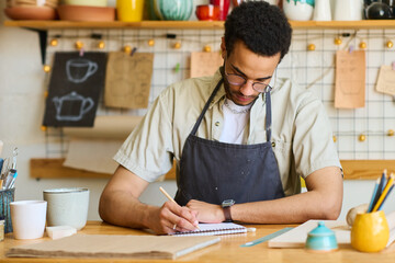 Aufkleber - African American guy in apron making notes in notepad while sitting by table and working over new collection of earthenware or clay items