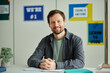 © Seventyfour - Front view portrait of smiling bearded teacher looking at camera while sitting at desk in school classroom against colorful posters