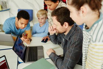 Wall Mural - High angle portrait of male teacher with diverse group of kids using laptop in school