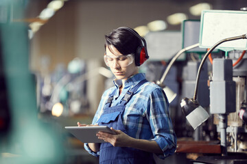 Wall Mural - Serious busy young female worker in ear protectors and safety goggles using digital tablet while making notes on processes of production shop