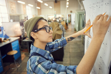 Wall Mural - Side view of focused young woman in glasses standing at factory shop and drafting technical sketch leaned to wall